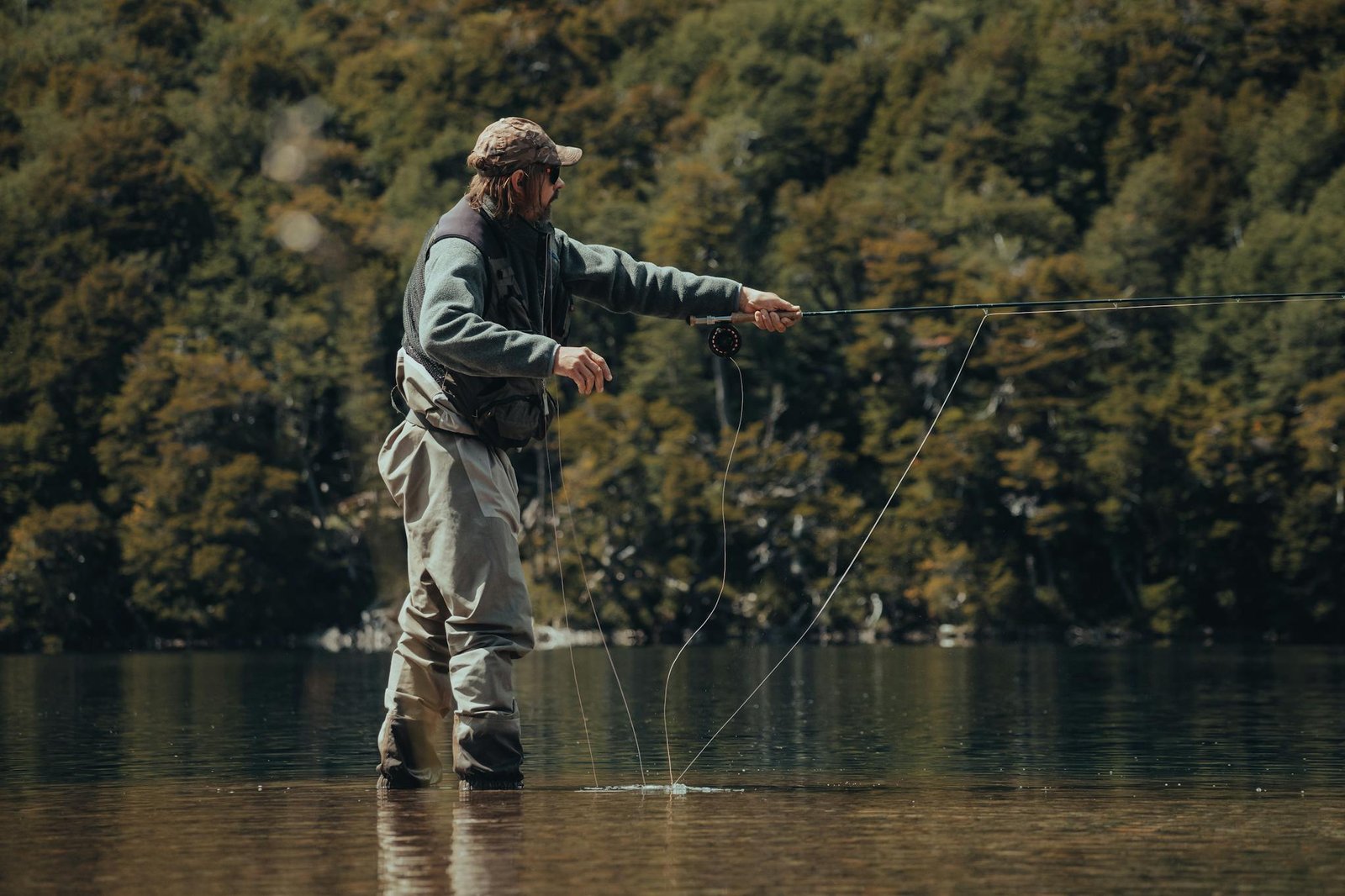 A man in waders fly fishing in a tranquil lake surrounded by lush green forest.